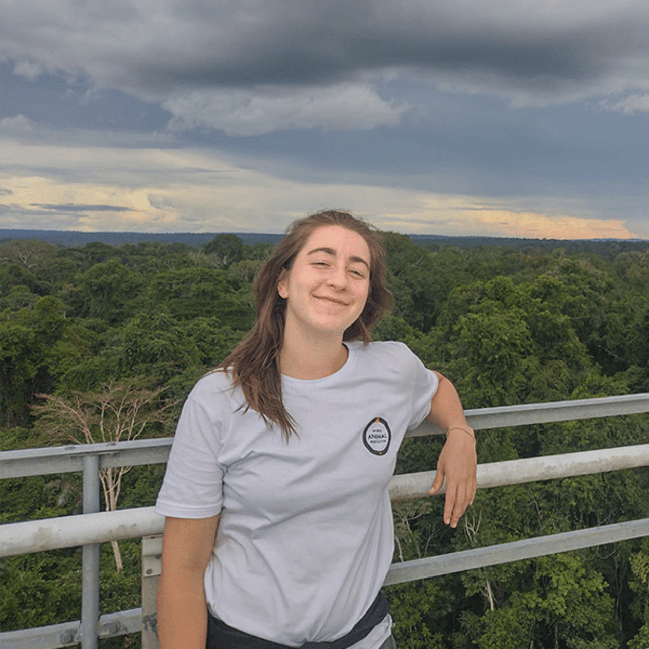 Júlia está em pé, se apoiando em uma grade, com uma grande vegetação ao fundo