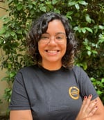 Natália Figueiredo é uma mulher de cabelos curtos e cacheados, ela está olhando para a câmera, sorrindo e com os braços cruzados.