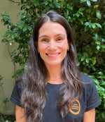 Marina é uma mulher branca de cabelos castanhos escuros. Ela está olhando pra frente e sorrindo.