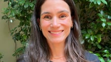 Marina é uma mulher branca de cabelos castanhos escuros. Ela está olhando pra frente e sorrindo.