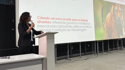Natália Figueiredo, atrás de um púlpito, durante o evento na Universidade Federal da Paraíba. Natália segura um microfone nas mãos e atrás dela tem um telão com uma apresentação sobre o seu trabalho na Proteção Animal Mundial.
