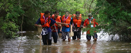 Grupo de cinco homens do movimento Escoteiros do Brasil, com coletes salva-vidas, andando em um rio que bate em seus joelhos.